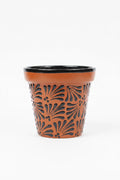 A terracotta-colored ceramic planter pot with a black rim and a black floral cutout pattern on the front, hand painted and made in Mexico.