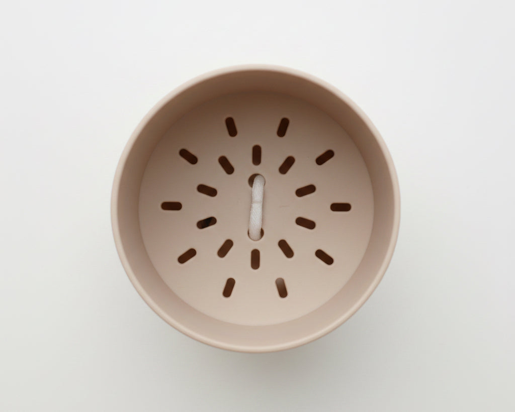 A top-down view of a light beige plastic cup insert or lid featuring a circular pattern of small cutouts and a small white central tab, set against a textured gray background.